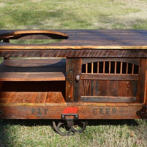 May include: A rustic wooden cart with a built-in grill and a cabinet door. The cart has wheels and is painted with the words "PIT" and "CREW".