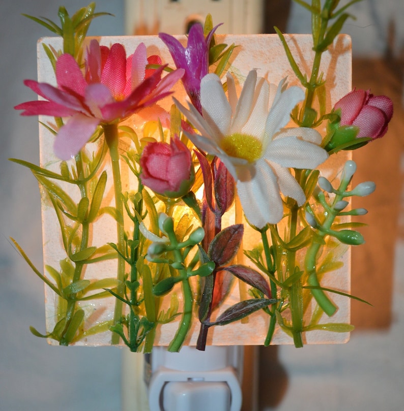 May include: A white night light with a square glass panel featuring a floral arrangement of pink, white, and purple flowers with green leaves. The light is plugged into an outlet.