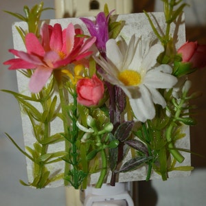May include: A white nightlight with a floral design. The flowers are pink, white, and yellow. The nightlight is plugged into an outlet.