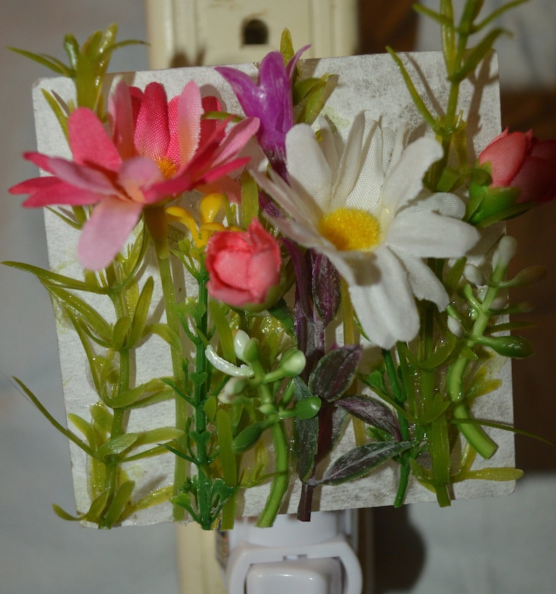 May include: A white nightlight with a floral design. The flowers are pink, white, and yellow. The nightlight is plugged into an outlet.