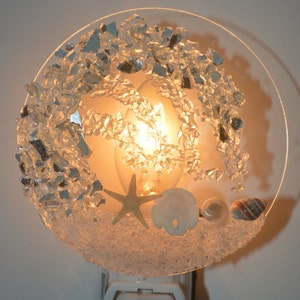 May include: A decorative night light with a beach theme. The round, clear plastic base features a warm light, surrounded by crushed glass, a starfish, a sand dollar, a snail shell, and a striped seashell. The night light is plugged into a white outlet.