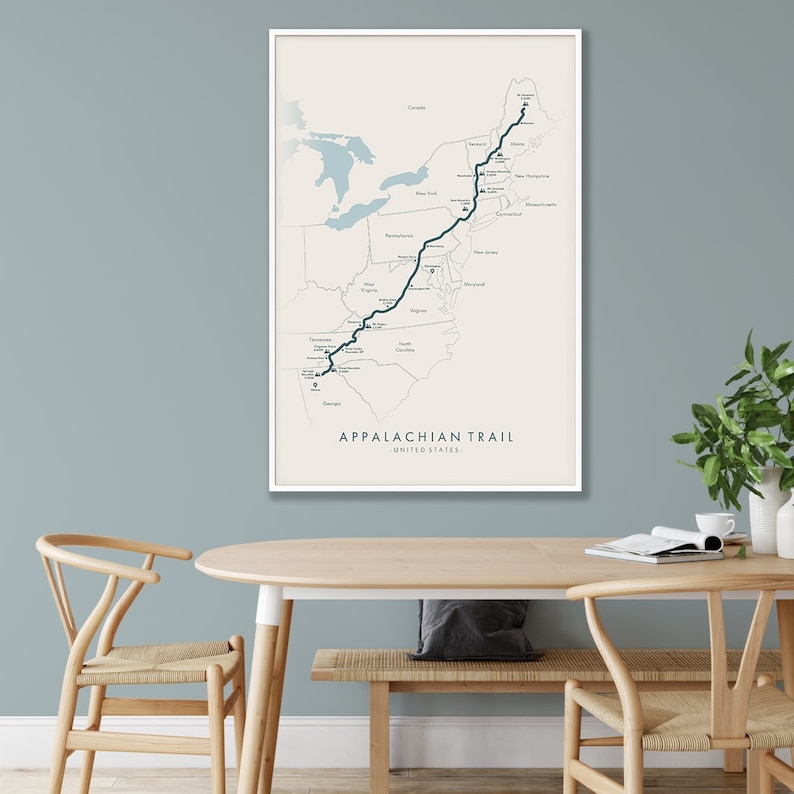 May include: A beige map print with a dark blue line tracing the Appalachian Trail across the United States. The map is framed in a white frame and hanging on a light blue wall.
