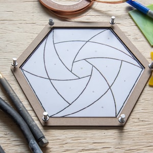 May include: A hexagonal wooden frame holds a white paper design with black lines and lettered sections, possibly for stained glass. Pliers, a glass cutter, and a Sharpie marker are present, alongside copper foil and coloured glass pieces.