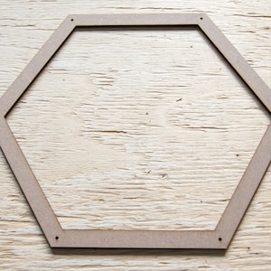 May include: An unfinished wooden hexagon frame in a light brown hue, featuring six sides and small pre-drilled holes at each corner. The frame is placed on a light-coloured wooden surface, highlighting the natural wood grain.