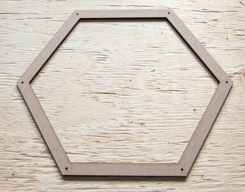 May include: An unfinished wooden hexagon frame in a light brown hue, featuring six sides and small pre-drilled holes at each corner. The frame is placed on a light-coloured wooden surface, highlighting the natural wood grain.
