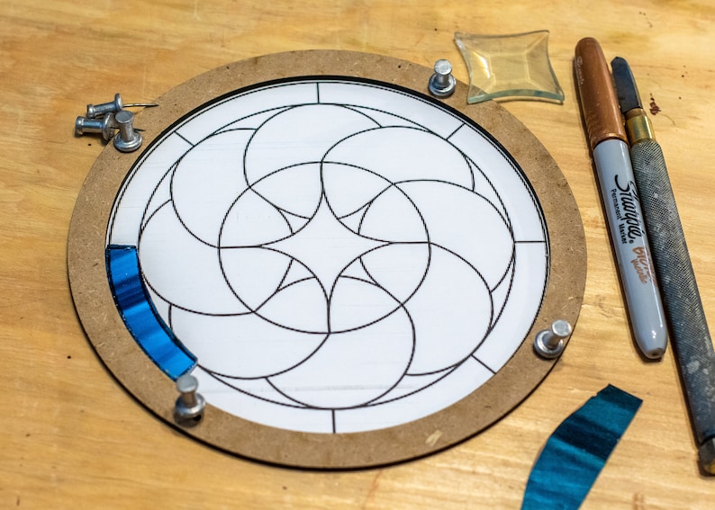 May include: A circular stained glass project in progress, featuring a white paper pattern with black outlines, secured within a wooden frame. Blue glass pieces are partially placed. Tools include a Sharpie marker and a glass cutter.