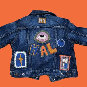Può includere: Una giacca di jeans blu con varie toppe colorate, tra cui un grande occhio, la parola "HALO", una stella e una finestra. La giacca ha una piccola toppa "NY" sul retro.