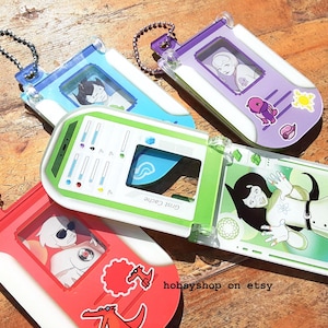 Puede incluir: Cuatro dispositivos electrónicos de juegos portátiles de colores con gráficos de personajes de dibujos animados. Los dispositivos son azules, morados, verdes y rojos, cada uno con un diseño diferente y una cadena adjunta. El dispositivo verde tiene el texto "Grist Cache".