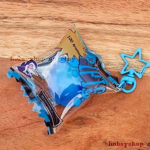 Könnte beinhalten: Ein blau-schwarzer Schlüsselanhänger mit einem sternförmigen Verschluss. Der Schlüsselanhänger ist an einer transparenten Plastikverpackung befestigt, die eine blaue Kugel enthält. Die Verpackung hat blaue und schwarze Akzente und den Text "100 Kronen".