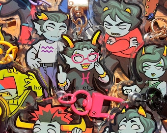 May include: A collection of cartoon character keychains with various designs. The keychains feature characters with gray skin, horns, and unique outfits. Some keychains have pink accents, and the text "hopsyshop on etsy" is visible.