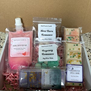 May include: A gift box containing soy wax melts and a squeezable wax pouch. The box includes wax melts in various colours and scents, such as "Book Boyfriend," "Slow Burn Romance," and "Enchanted Spell Book." The pouch is pink and the box is lined with pink paper.