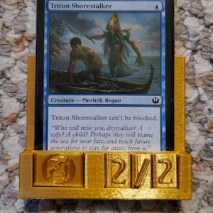 Puede incluir: Una carta azul y blanca de Magic: The Gathering con una imagen de una criatura con capucha y una figura humana en un bote. El texto de la carta dice "Triton Shorestalker" y "Criatura - Merfolk Rogue". La carta tiene una fuerza de 2 y una resistencia de 2.