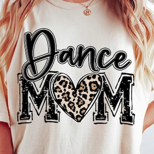 May include: A white t-shirt with the text "Dance Mom" in black with a leopard print heart in the middle of the word "Mom". The shirt is being worn by a person with long blonde hair. The text "Digital Download PNG only" is in a pink circle with a white outline.