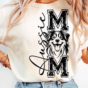 May include: A white t-shirt with a black graphic design featuring a dog's head and the text "Aussie Mom" in a stylized font.
