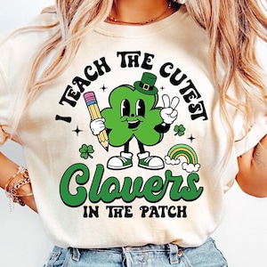 May include: A white t-shirt with a green shamrock cartoon character holding a pencil and a rainbow. The text on the shirt reads "I Teach The Cutest Clovers In The Patch".