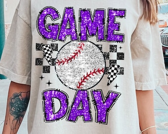 May include: A white t-shirt with a purple glitter design that says "GAME DAY" with a baseball in the middle. The baseball has white stitching and a red seam. The design is surrounded by black and white checkered flags.