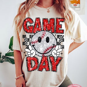 Puede incluir: Una camiseta blanca con un diseño de purpurina roja que dice "GAME DAY" con un gráfico de béisbol sonriente en el centro. El diseño también incluye un patrón a cuadros y un rayo.