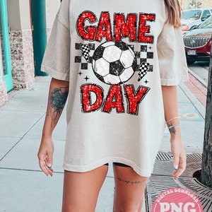 May include: A white t-shirt with a red glitter graphic that says "GAME DAY" with a soccer ball and checkered flags.