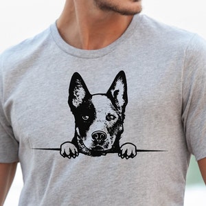 May include: A gray t-shirt featuring a black and white graphic of a dog's head and paws peeking over a horizontal line. The image includes the text "SVG, PNG, DXF, EPS" in the upper left corner. The person is wearing blue jeans.