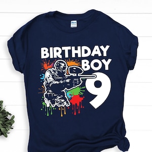 Op de afbeelding: Een marineblauw t-shirt met een wit ontwerp van een paintballspeler met kleurrijke verfspatten en de tekst "Birthday Boy 9".