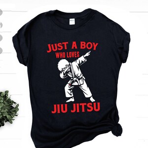 Puede incluir: Camiseta negra con texto blanco que dice "Just a boy who loves Jiu Jitsu". Un dibujo animado de un niño con un gi blanco haciendo un dab está por encima del texto.