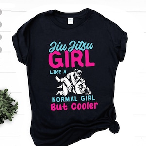 Puede incluir: Camiseta negra con un gráfico de una mujer en un combate de jiu-jitsu. El texto en la camiseta dice "Jiu Jitsu Girl Like A Normal Girl But Cooler".