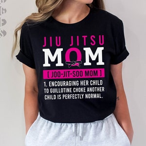 Może przedstawiać: Czarny t-shirt z napisem "JIU JITSU MOM" w kolorze białym i różowym. Projekt zawiera grafikę czarnego paska, frazę "[JOO-JIT-SOO MOM]" i humorystyczny cytat o rodzicielstwie.
