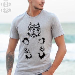 May include: A gray t-shirt featuring a black graphic of a dog's head and paws bursting through the fabric. The shirt has the text "SVG, PNG, DXF, EPS" in the upper left corner. The person is wearing blue jeans.