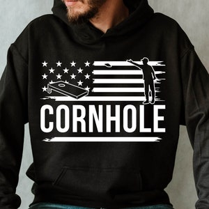以下が含まれることがあります： 白いグラフィックでコーンホールボード、星、ストライプ、豆袋を投げている人を示す黒いパーカー。 「CORNHOLE」の文字は白で印刷されています。 パーカーは木製のスツールに座っている人が着用しています。