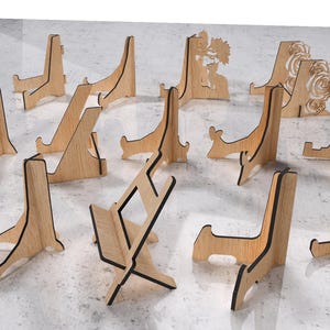 May include: A collection of wooden display stands in various designs. Some stands feature cut-out butterfly, floral, and geometric patterns. The stands are made of light-colored wood with dark edges and are suitable for displaying items.