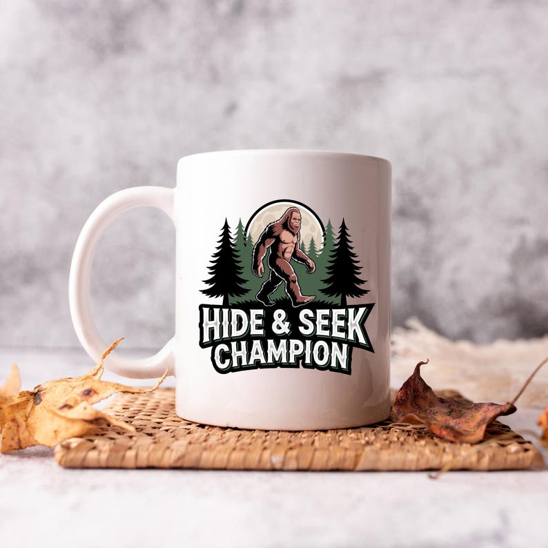 Puede incluir: Taza de cer&aacute;mica blanca con un gr&aacute;fico de Bigfoot. El dise&ntilde;o presenta un Bigfoot caminando frente a una luna llena y pinos, con el texto "HIDE & SEEK CHAMPION" en fuente verde y blanca. La taza tiene un asa curva.