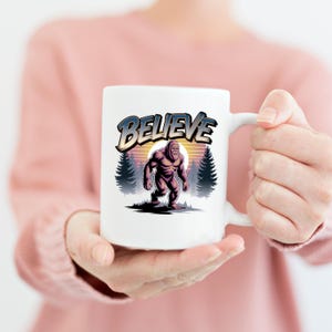 Puede incluir: Taza de cer&aacute;mica blanca con una ilustraci&oacute;n de Bigfoot y la palabra "BELIEVE". El dise&ntilde;o incluye un fondo de puesta de sol y pinos, creando una est&eacute;tica vintage. La taza es sostenida por una persona.