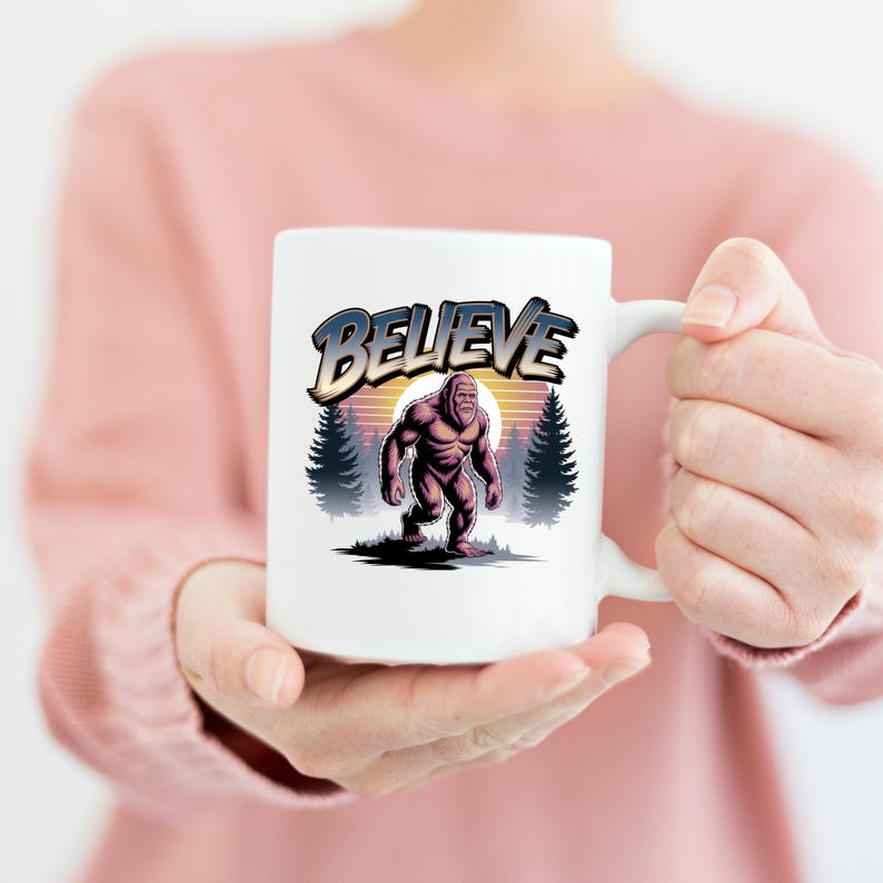 Puede incluir: Taza de cer&aacute;mica blanca con una ilustraci&oacute;n de Bigfoot y la palabra "BELIEVE". El dise&ntilde;o incluye un fondo de puesta de sol y pinos, creando una est&eacute;tica vintage. La taza es sostenida por una persona.