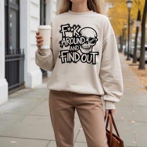 K&ouml;nnte beinhalten: Beiges Sweatshirt mit dem Text "F**k Around and Find Out" in schwarzer, verwaschener Schrift, mit einem Totenkopf mit Sonnenbrille. Das Sweatshirt wird mit einer braunen Hose kombiniert.