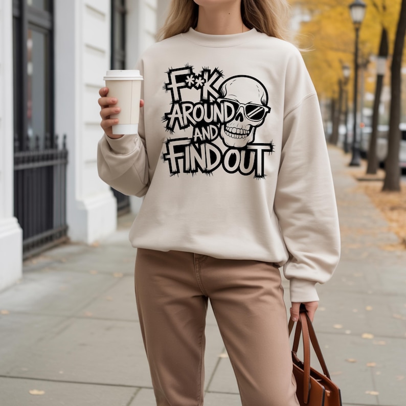 K&ouml;nnte beinhalten: Beiges Sweatshirt mit dem Text "F**k Around and Find Out" in schwarzer, verwaschener Schrift, mit einem Totenkopf mit Sonnenbrille. Das Sweatshirt wird mit einer braunen Hose kombiniert.