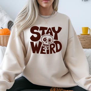 K&ouml;nnte beinhalten: Beiges Sweatshirt mit dem Schriftzug "STAY WEIRD" in dunkelbraunen Buchstaben. Das Wort "WEIRD" zeigt einen Cartoon-Sch&auml;del mit herausgestreckter Zunge. Das Sweatshirt ist ein l&auml;ssiger, bequemer Stil.