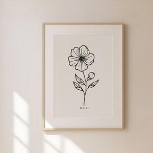May include: A black and white line drawing of a flower with a single stem and leaves. The drawing is framed in a light wood frame and is hanging on a white wall.