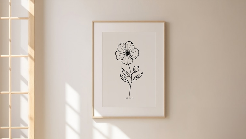 May include: A black and white line drawing of a flower with a single stem and leaves. The drawing is framed in a light wood frame and is hanging on a white wall.