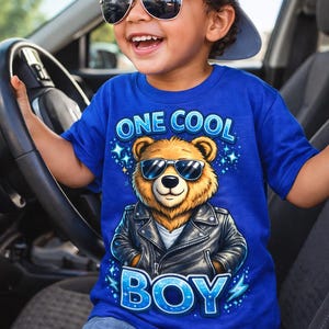 May include: A young child wearing a blue t-shirt with the text "ONE COOL BOY" and a graphic of a bear wearing sunglasses and a leather jacket. The child is also wearing sunglasses and a gray baseball cap.