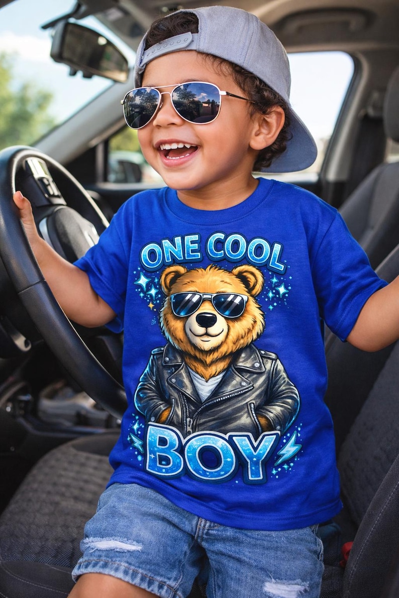 May include: A young child wearing a blue t-shirt with the text "ONE COOL BOY" and a graphic of a bear wearing sunglasses and a leather jacket. The child is also wearing sunglasses and a gray baseball cap.
