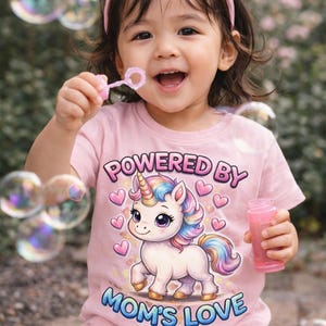 May include: A young child wearing a pink t-shirt with a unicorn graphic and the text "POWERED BY MOM'S LOVE". The child is smiling and blowing bubbles with a pink wand. A pink headband is in the child's hair.