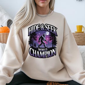 Puede incluir: Sudadera beige con un gr&aacute;fico que presenta una ilustraci&oacute;n de Bigfoot caminando en un bosque bajo un cielo estrellado y el texto "HIDE & SEEK WORLD CHAMPION". El dise&ntilde;o incluye acentos morados y blancos.