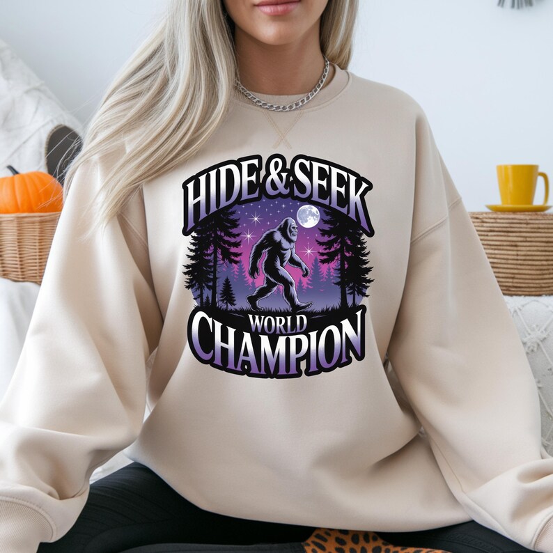 Puede incluir: Sudadera beige con un gr&aacute;fico que presenta una ilustraci&oacute;n de Bigfoot caminando en un bosque bajo un cielo estrellado y el texto "HIDE & SEEK WORLD CHAMPION". El dise&ntilde;o incluye acentos morados y blancos.