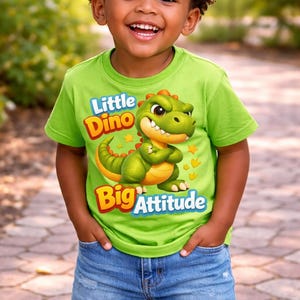 May include: A young child smiles, wearing a lime green t-shirt with a cartoon dinosaur graphic. The shirt reads "Little Dino Big Attitude" in yellow and red text. The child is also wearing blue denim shorts.