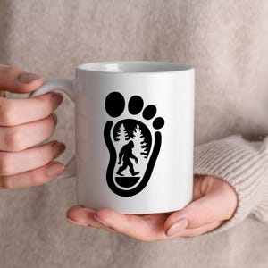 May include: White ceramic mug with a black silhouette of a Bigfoot footprint. Inside the footprint is a silhouette of Bigfoot walking through a forest.