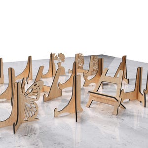 May include: A collection of wooden display stands in various designs, including a butterfly, floral, and geometric patterns. The stands are made of light-colored wood and are arranged on a light-colored surface.