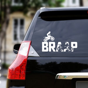 Op de afbeelding: Een witte vinyl sticker met een silhouet van een persoon die een crossmotor rijdt en het woord "BRAAP" op een zwarte auto.