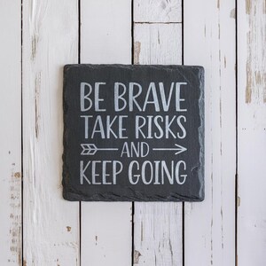 May include: Black slate sign with white text that reads "Be Brave Take Risks And Keep Going" with a white arrow pointing to the right.