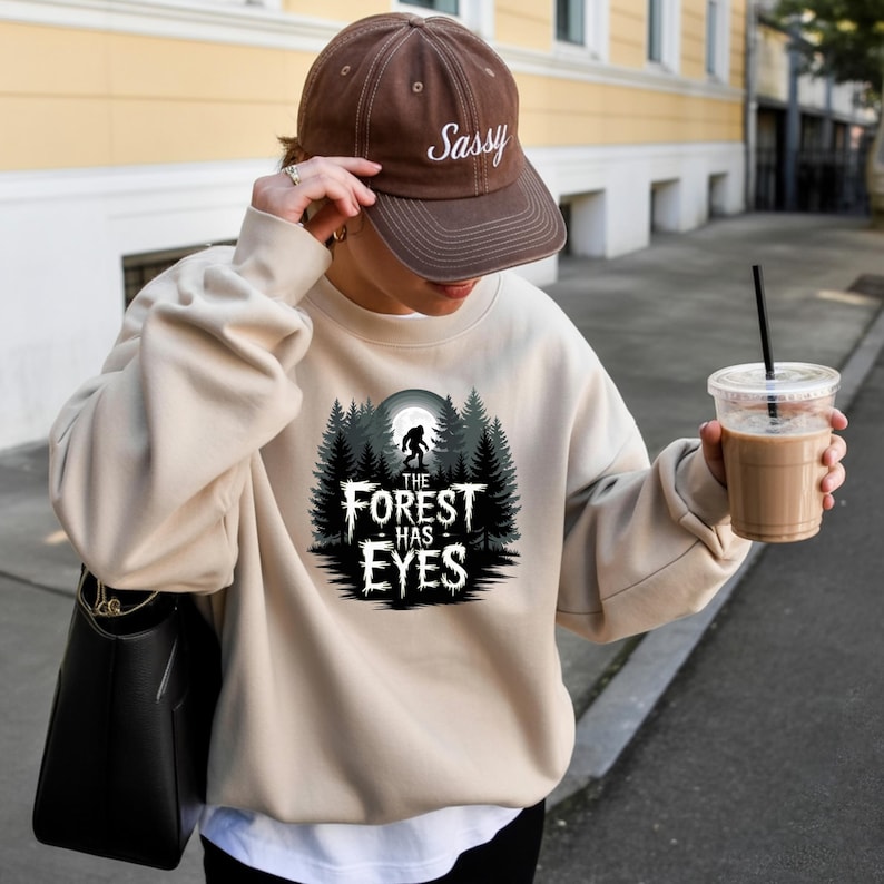 Puede incluir: Sudadera beige con un gr&aacute;fico de un bosque y el texto "THE FOREST HAS EYES". La sudadera se combina con una gorra de b&eacute;isbol marr&oacute;n que dice "Sassy". Tambi&eacute;n se ven un bolso negro y una bebida de caf&eacute;.