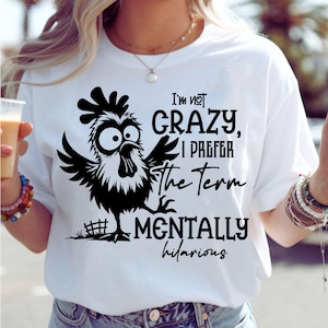 Może przedstawiać: Biała koszulka z czarnym rysunkowym kogutem i napisem "I'm not crazy, I prefer the term mentally hilarious." Kogut ma duże oczy i rozłożone skrzydła. Koszulka jest w stylu casual.
